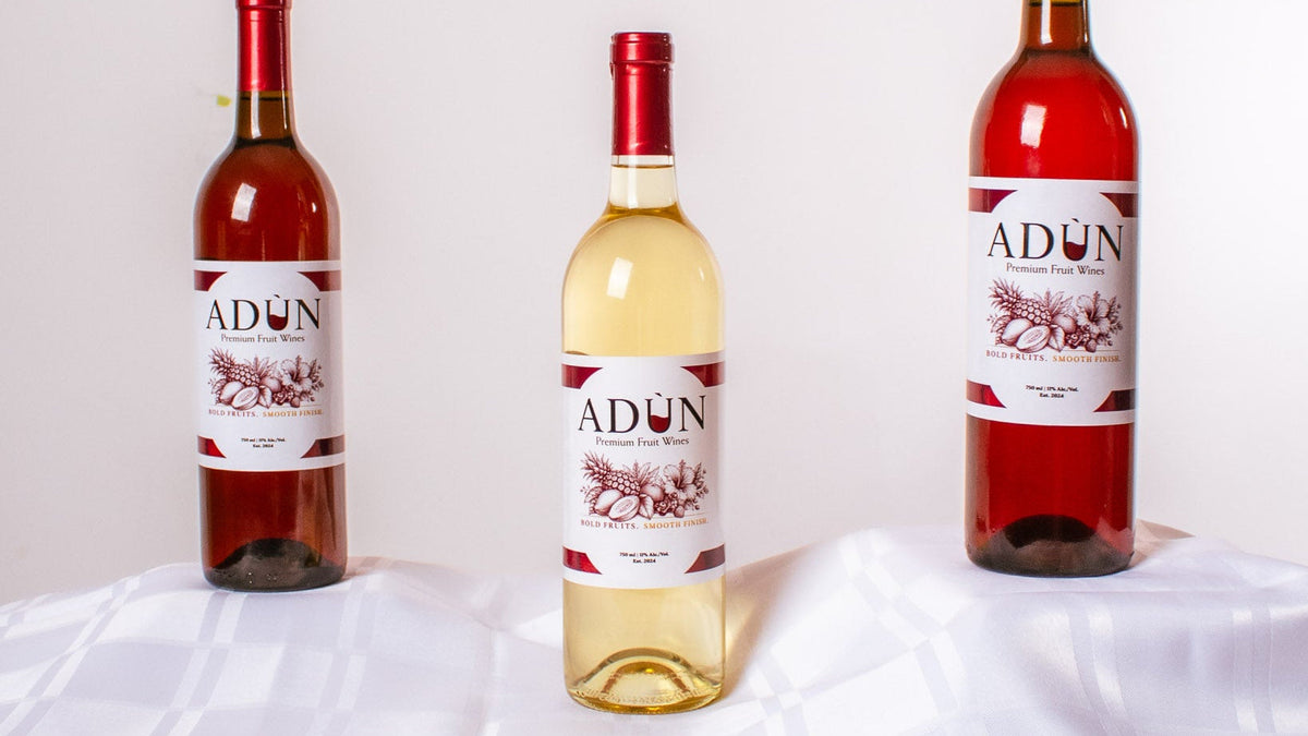 Three bottles of ADUN wine arranged on a white draped table. The left bottle contains a deep red wine, the center bottle is a white wine, and the right bottle contains a rosé tone wine.