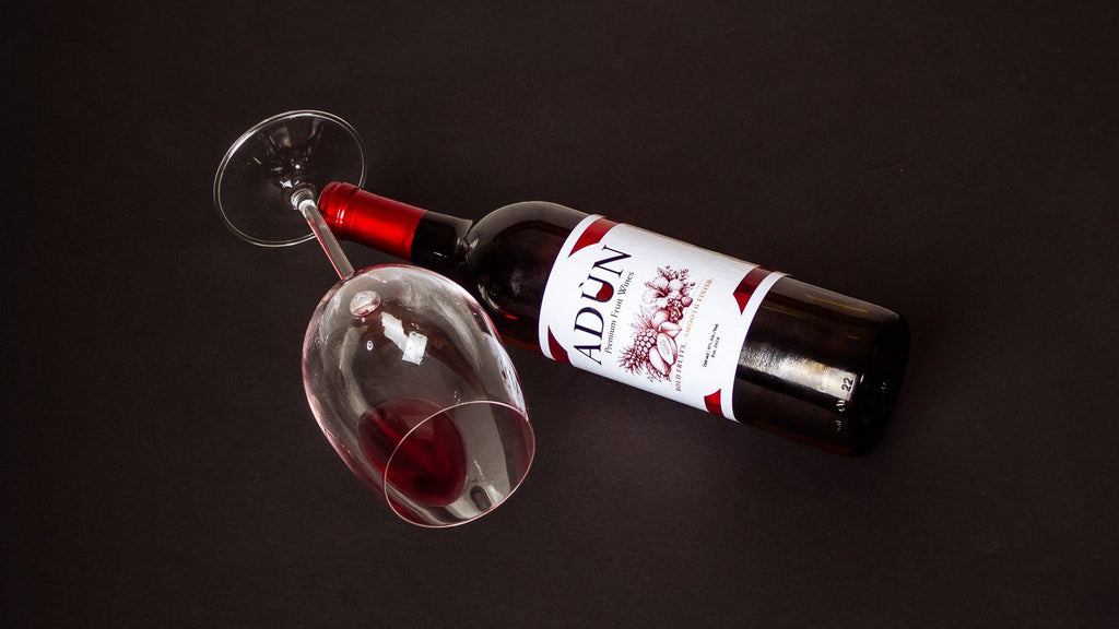 A bottle of red wine with a white and red label lies on its side next to a partially filled wine glass on a dark black background. The brand name 'Adùn' is visible on the label.