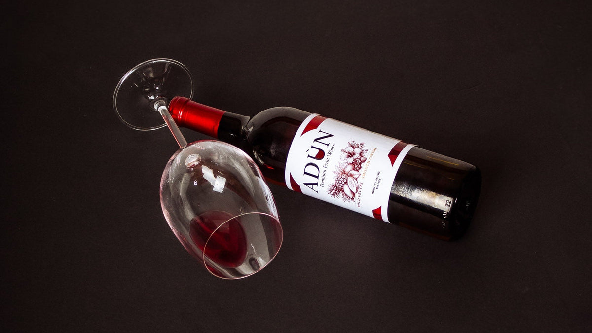 A bottle of red wine with a white and red label lies on its side next to a partially filled wine glass on a dark black background. The brand name 'Adùn' is visible on the label.