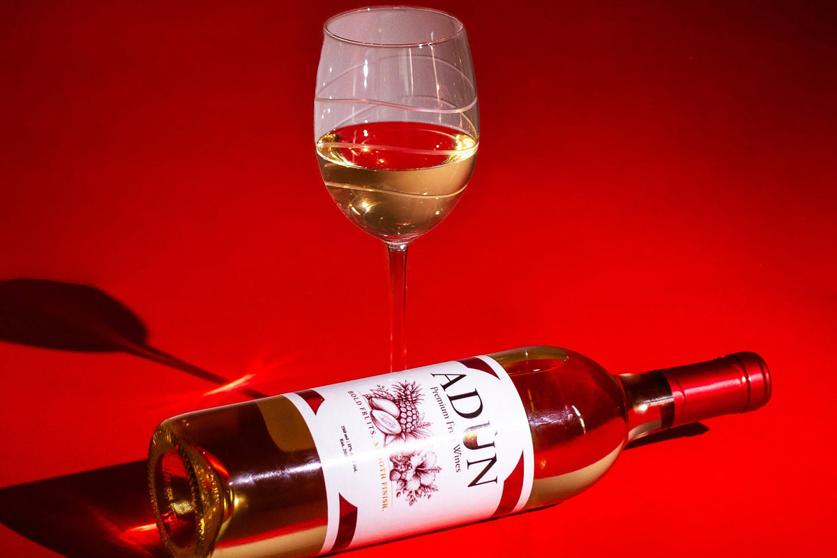 A bottle of white wine lies on its side next to a filled wine glass against a vivid red background. The wine bottle label shows the brand name 'Adun'.